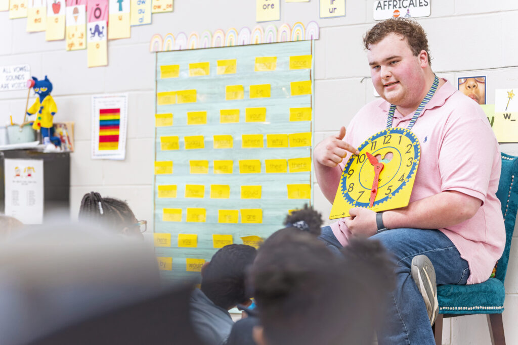 A young man holds a cardboard clock in front of a room full of students