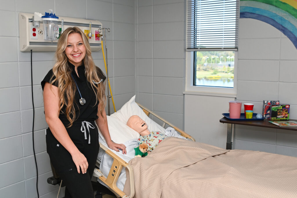 A young nursing instructor stands next to a hospital bed with a pediatric manikin in it